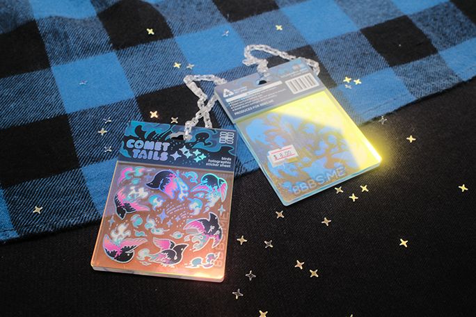 Photo of 2 acrylic charms styled to look like packaged sticker sheets. They are printed on rainbow acrylic with holographic silver foil accents, are themed on birds and titled "Comet Tails". The back of the fake packaging header contains some plausible usage warnings and advice and a barcode, and a fake price tag displays the cost to be $8.00 (not the actual charm's cost). The charms are laid on a cloth backdrop with scattered four-pointed star glitter.