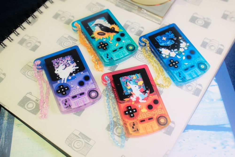Four colourful acrylic charms shaped like game boys, arranged on a large notebook. The print is layered to show depth. Each charm features a bunny or crow together with a plant  for each of the seasons. 