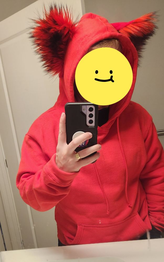 A mirror selfie of a white man, at his bathroom counter. He is wearing a red hoodie, which has a set of red ears on the hood. His face is obscured by a smiley face.