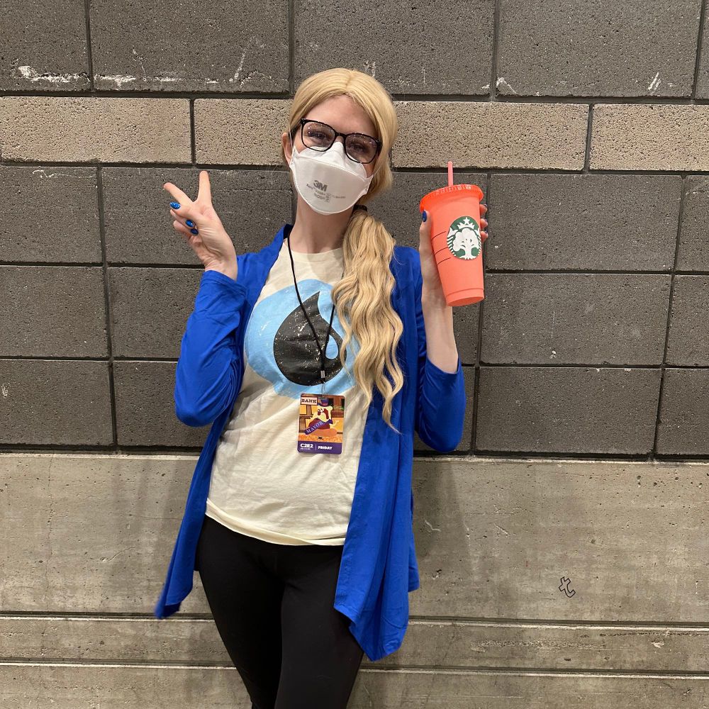 Woman in the color scheme of an island mana in magic the gathering, with Ugg slippers, black leggings, a blue mana shirt, a blue cardigan, a blonde wig, and Starbucks cup with a green mana logo on the front