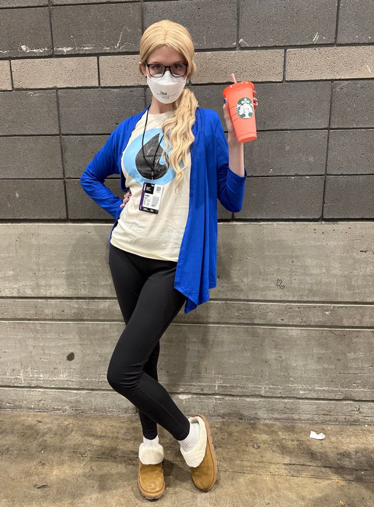 Woman in the color scheme of an island mana in magic the gathering, with Ugg slippers, black leggings, a blue mana shirt, a blue cardigan, a blonde wig, and Starbucks cup with a green mana logo on the front