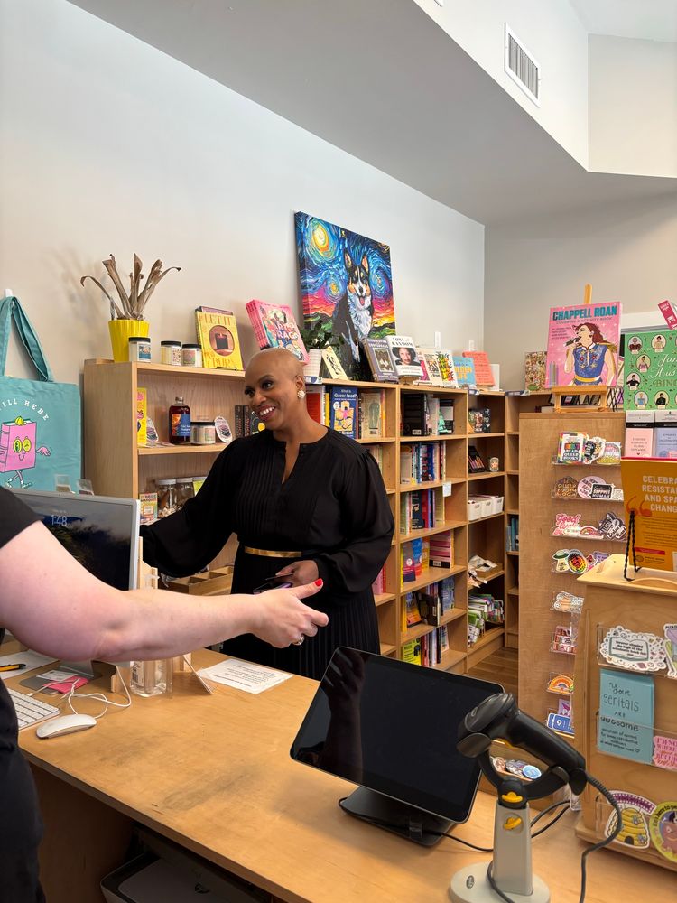 Rep. Pressley, wearing a black dress, smiles as she checks out a book. A woman’s hand reaches out for her book across the check out counter. Behind them is the bookstore, filled with colorful art and books.