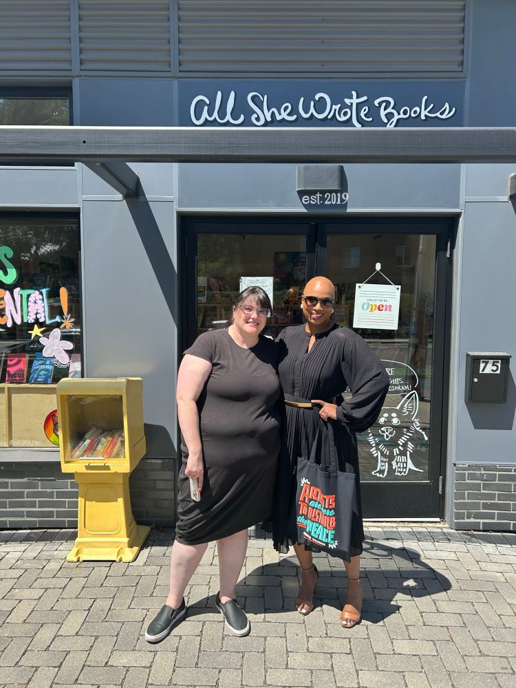 Rep. Pressley, in a black dress and black tote bag, poses next to a woman in a black dress in front of the bookstore front door. The bookstore is called “All She Wrote Books.”