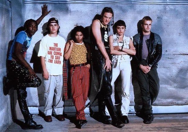A cast photo of HACKERS (1995) where The Phantom Phreak is actually present. They're leaning against a wall covered in netting and kinda striking cool poses, vamping.