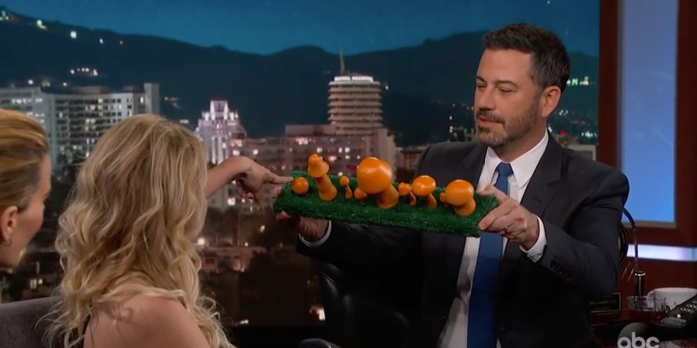 Photo. Jimmy Kimmel holds up a green astro turf covered plank with an array of orange mushroom sculptures. Stormy Daniels is pointing to a very short one.