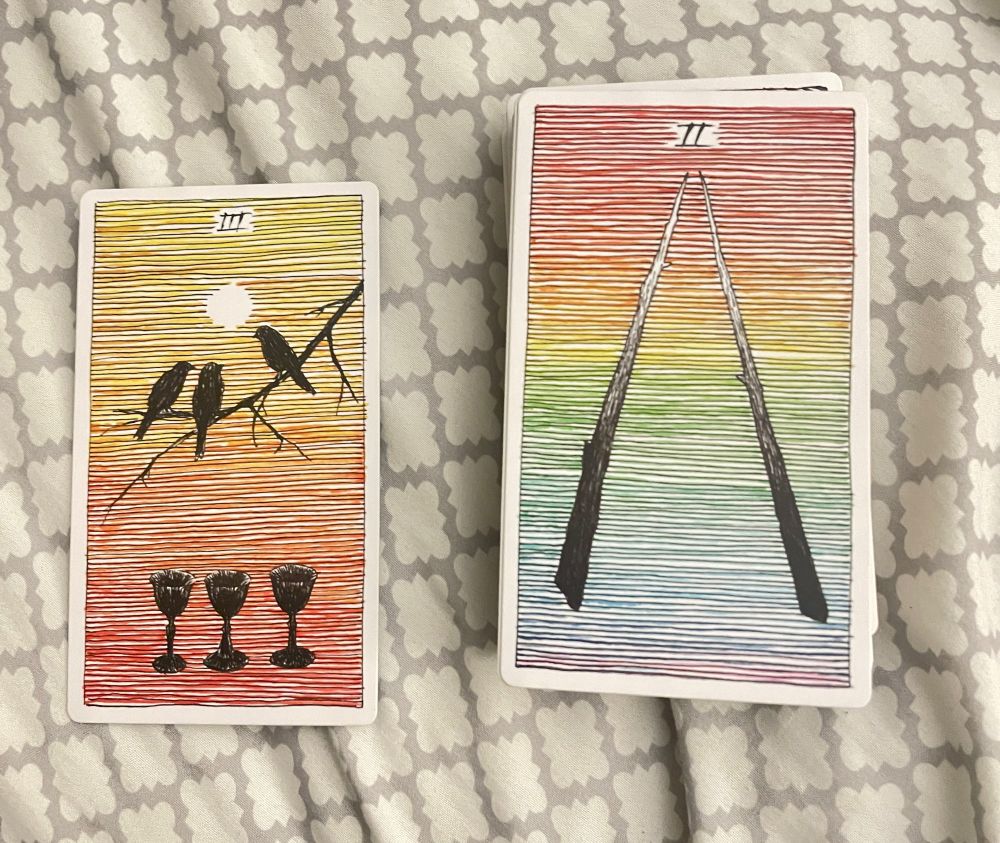 One card showing the roman numeral three and three birds on a branch over three cups with a sun in the background. 

Another card showing the roman numeral two and two sticks pointing upward with a rainbow gradient background.  