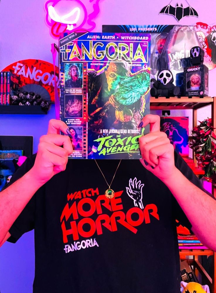 Fango's Social Media Coordinator LT (aka queerdem0n) holds the subscriber-exclusive cover over their face while wearing FANGORIA's WATCH MORE HORROR - EVIL THREADS tee.