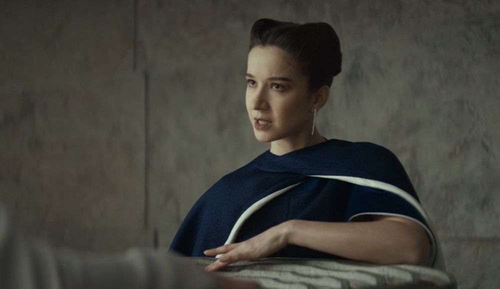 A screenshot from the Star Wars series Andor, showing an elegantly dressed, dark-haired woman reading a story on an ancient artifact through the touch of her fingers