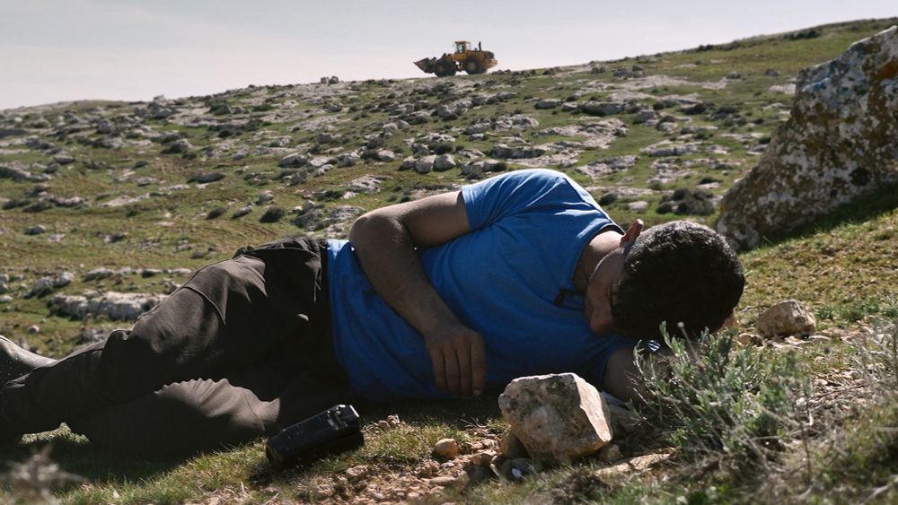 Plan du film. Badel Adra allongé sur ses terres, avec en arrière plan un bulldozer israélien 
