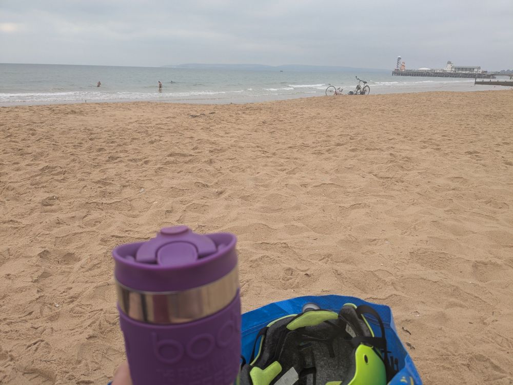 View of a sandy beach and the sea beyond, with a coffee cup and a bag containing a bike helmet in the foreground 
