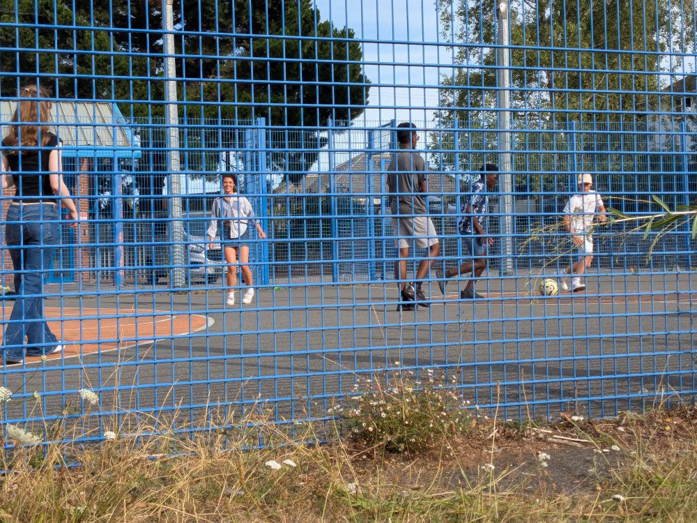 A photo through the blue mesh fence of a sports pitch, where a group of teenagers are playing football.