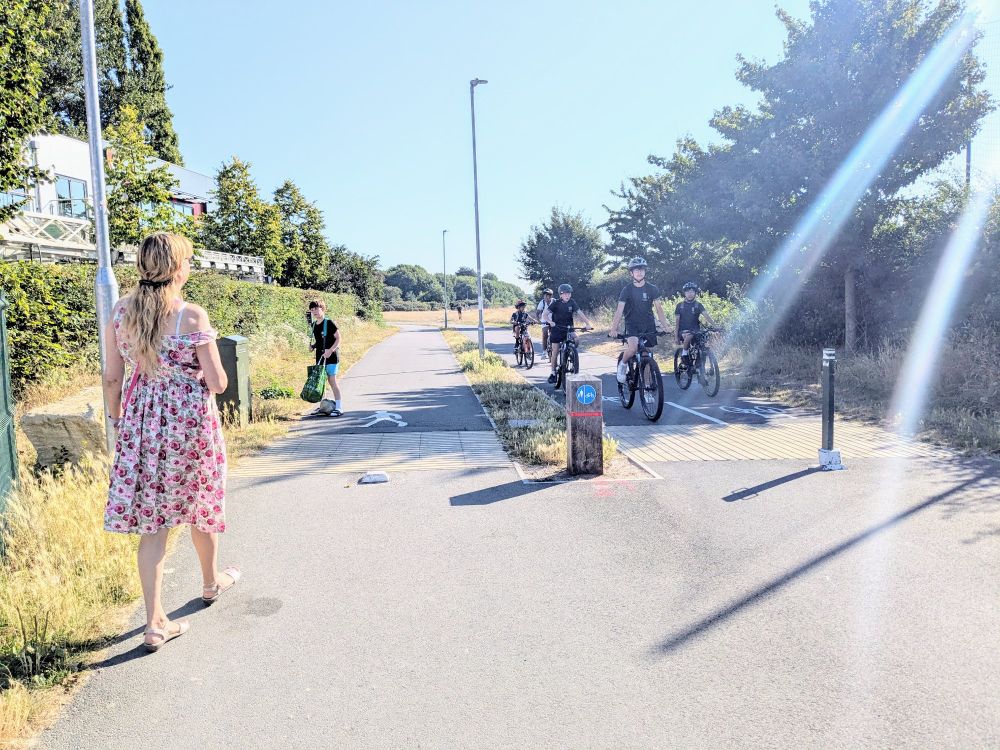 A segregated cycle path and footpath, with a lady and a child walking away from the camera, whilst a group of about six young teenagers in school uniform cycle in the opposite direction.