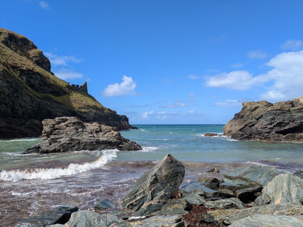 A stunning view looking out into a small rocky bay. The sky is a brilliant blue with just a few fluffy white clouds. The sea is choppy but a gorgeous green-blue colour.