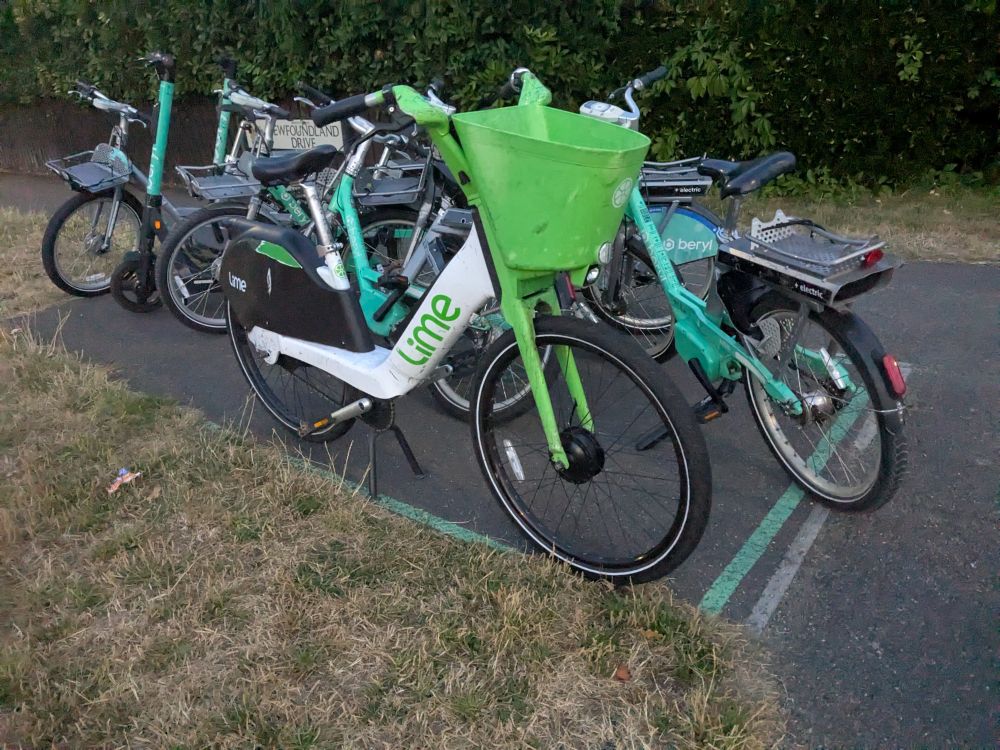Photo of several Beryl bike share bikes parked in a bay, along with one alien Lime bike.