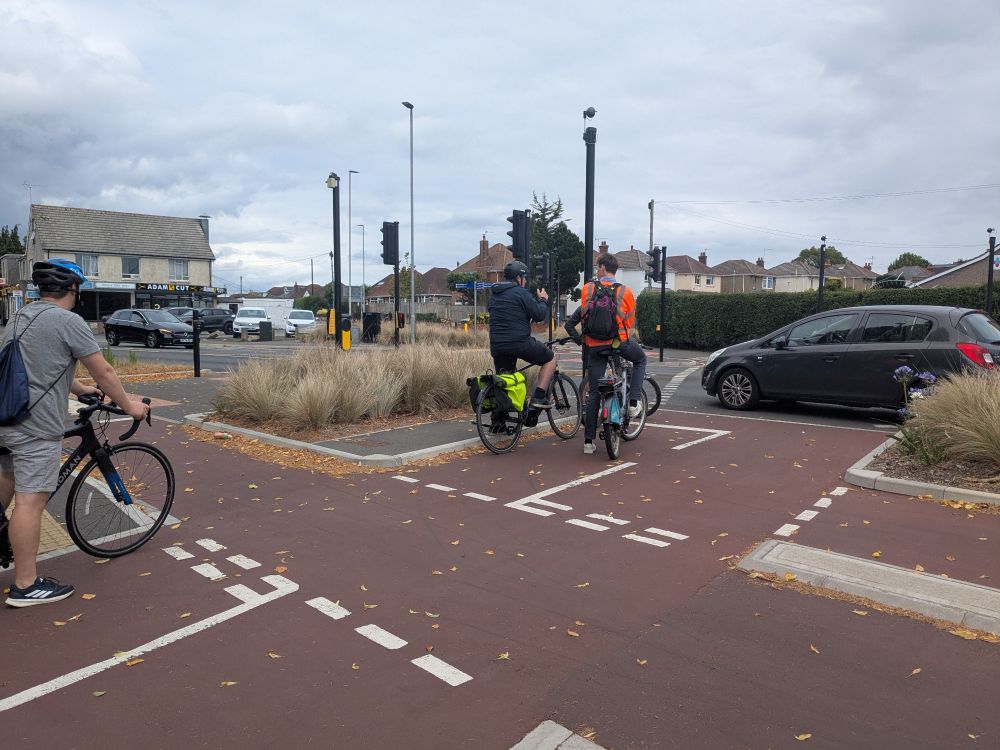 Three male cyclists at a complex road junction. The cycle path is paved in red tarmac and a car is passing through. Two of the cyclists are deep in conversation, probably talking about kerbs or signal phasing or something else rather geeky.