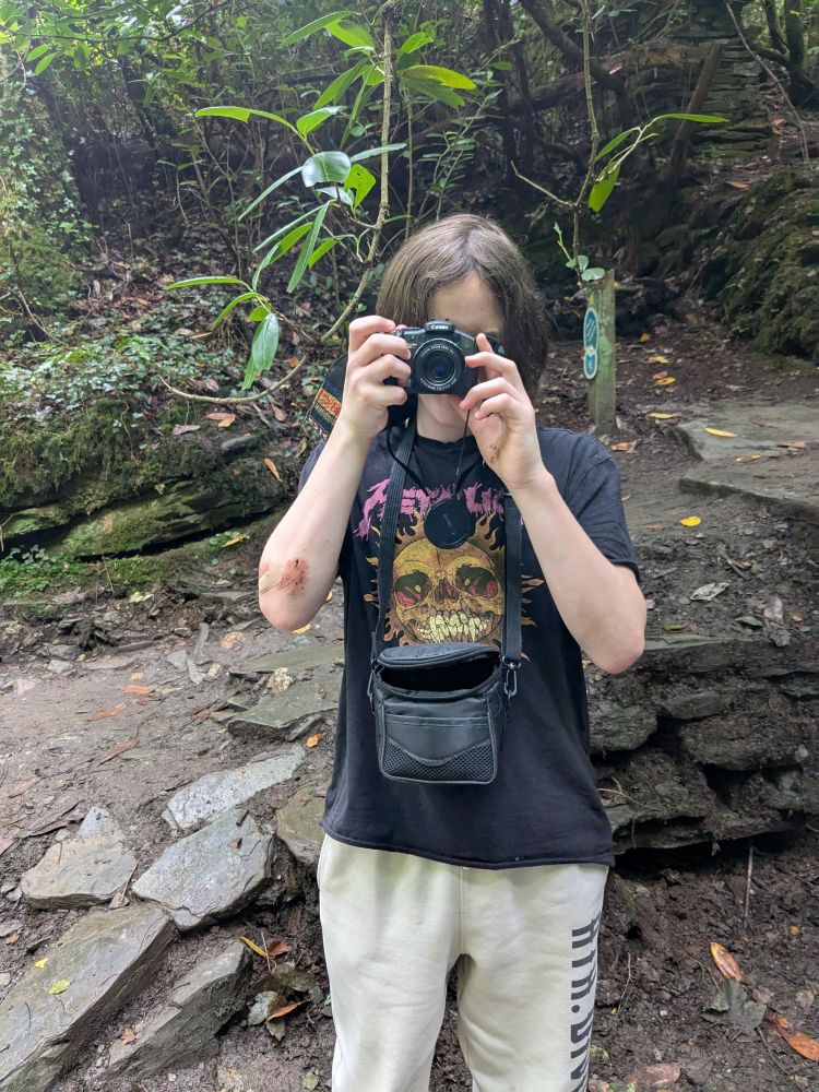 Photo of a thirteen year old boy taking a photo with a camera. He's wearing a Metallica tee shirt and has a nasty scrape on one elbow 