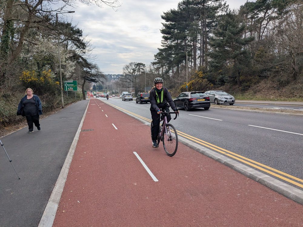 Photo of a segregated bi-directional cycle track with red surfacing alongside a dual carriageway. A smiling young woman is cycling towards the camera.