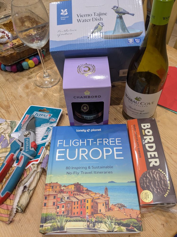 A selection of birthday presents laid out on a table: bottle of wine, bottle of Chambord, a bird feeder, garden secateurs, chocolate biscuits and a Lonely Planet book 'Flight Free Europe'