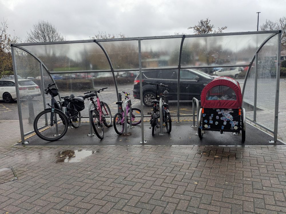 A bike shelter next to a car park, where a selection of bikes are parked, including some children's bikes and a bakfiets.