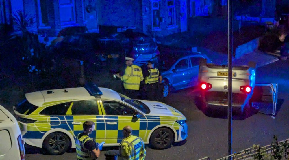 Photo of a dark residential street with a silver hatchback upside down, its lights still on, and a police car behind it. Several police officers are on the scene.