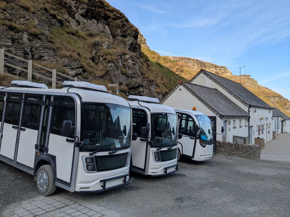 Three tiny electric buses that seat maybe 12 people or so, parked next to each other beside a cliff and ticket office at Tintagel.