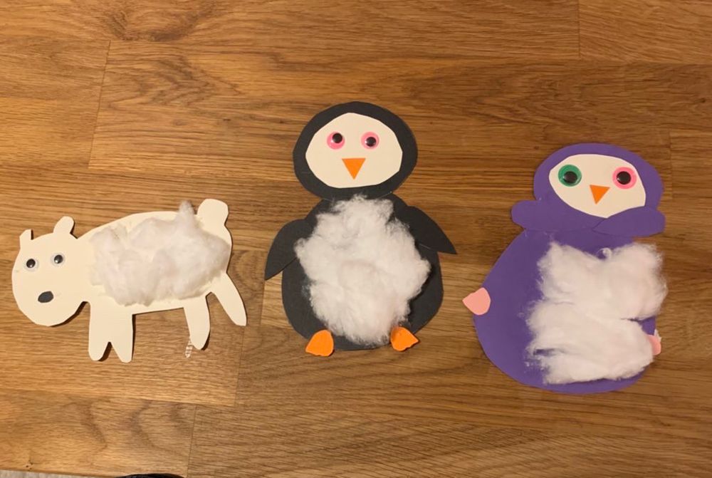 3 Tiere aus Tonkarton gebastelt auf einer Holzoberfläche. Das linke ist weiß mit weißen Wackelaugen, einem schwarzen Punkt als Nase und Watte als Fell. Eigentlich soll es ein Eisbär sein, sieht aber mehr aus wie ein Hund. In der Mitte ist ein Pinguin mit schwarzen Körper, mit weißem Gesicht und Watte auf dem Bauch. Dazu hat er einen orangen Schnabel und Füße und pinke Wackelaugen. Ganz rechts liegt noch ein Pinguin. Diesmal mit lila Körper. Das Gesicht ist wieder weiß und der Bauch mit Watte beklebt. Er hat einen orangen Schnabel und rosa Füßchen, die eher seitlich am Körper kleben. Er verschränkt die Flügel unterhalb des Gesichts und hat ein grünes Wackelauge (links) und ein pinkes Wackelauge (rechts).
