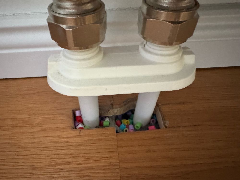 Zwei Heizungsrohre, die aus dem Boden herauskommen. Die Abdeckung zum Boden ist an den Rohren hochgeschoben. Darunter haben sich jede Menge bunte Bügelperlen angesammelt.