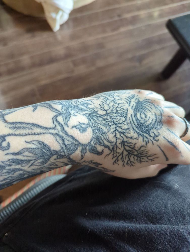 Photo of a man's left wrist and hand, showing a medieval style tattoo on the hand and wrist consisting of branches, leaves, the clawed feat of a monster, and a knot of wood that looks like an eye