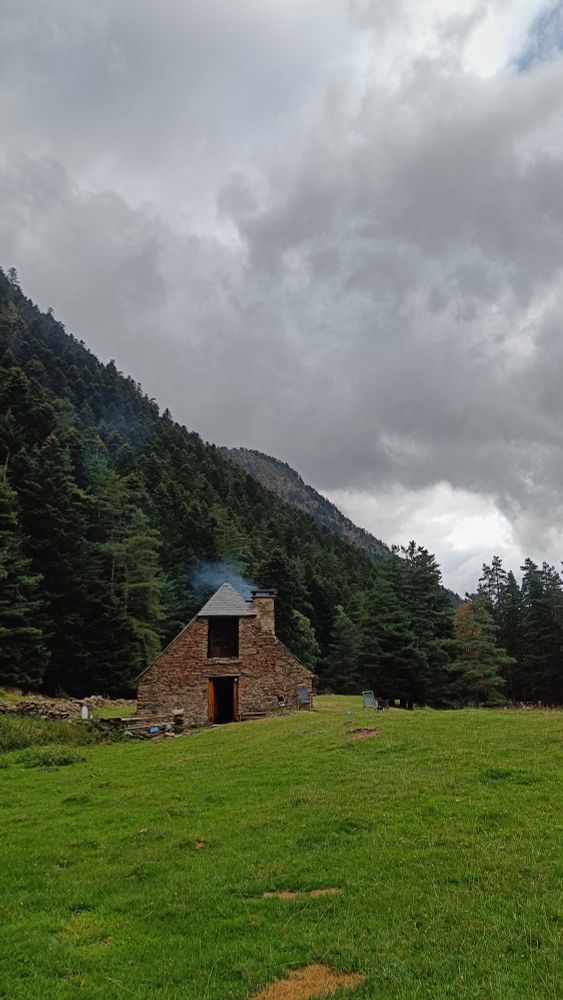 Une vieille grange pyrénéenne en pierre et toit d'ardoise au pied de montagnes. La cheminée fume.
