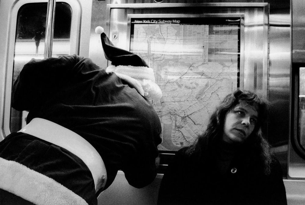 Black and white photo of a Santa in a NYC subway car leaning over a woman to look at the map. The woman's expression suggests she is bored, irritated, or just done. 