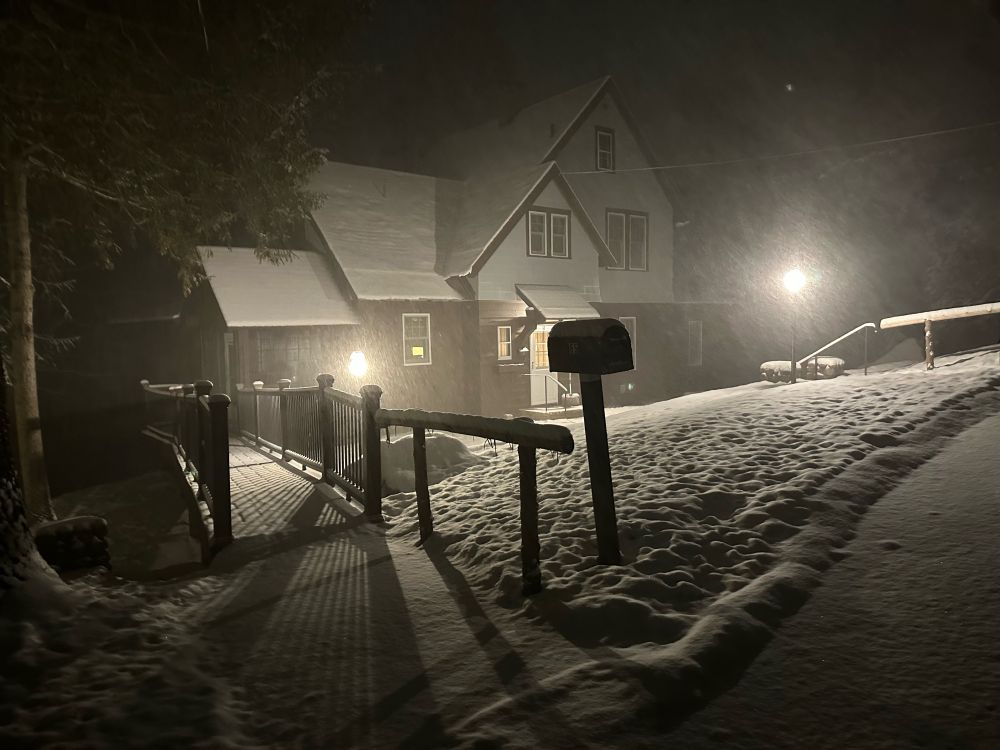 A house at night set back from the road illuminated with two standing lampposts, surrounded by snow on the ground and snow falling thickly