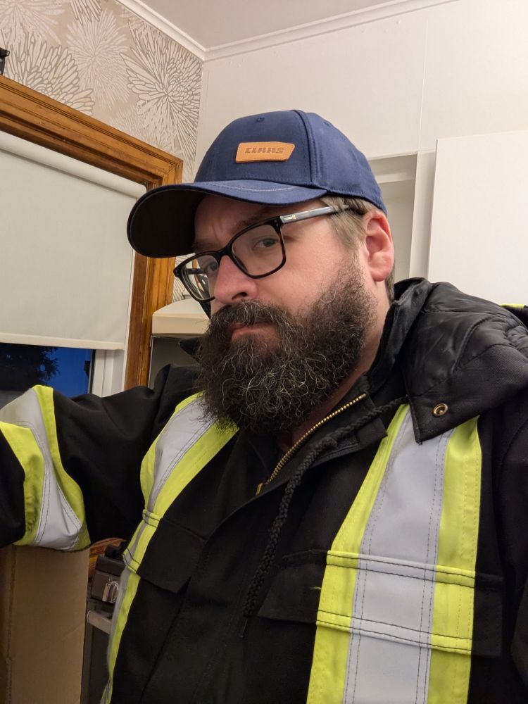 A bearded man in a black hi-vis jacket and a navy blue hat. The hat has a little brown leather patch that says "Claas."