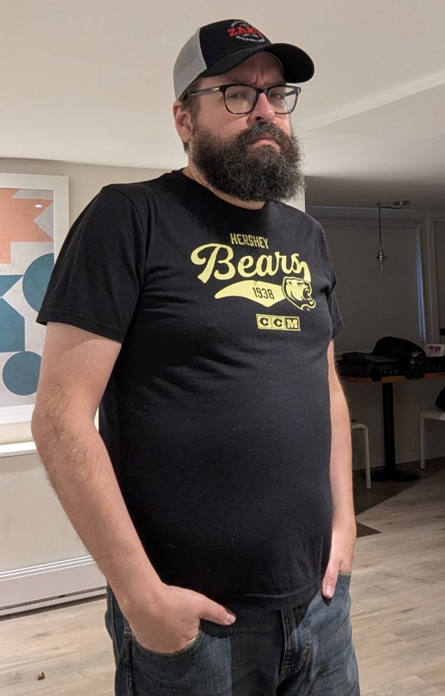 A bearded man in a black and grey hat that says "Zak's" and a black shirt that says "Hershey Bears 1938 CCM".