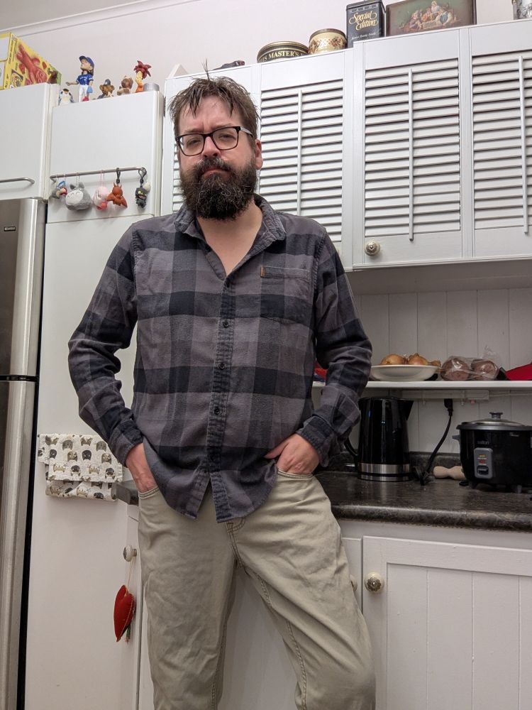 Bearded man in a black and grey flannel shirt and beige pants.