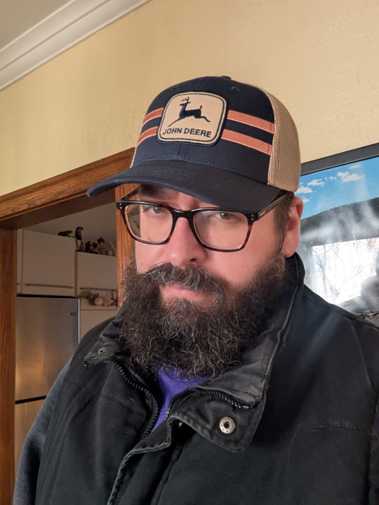 A bearded man wearing a blue and brown John Deere hat.