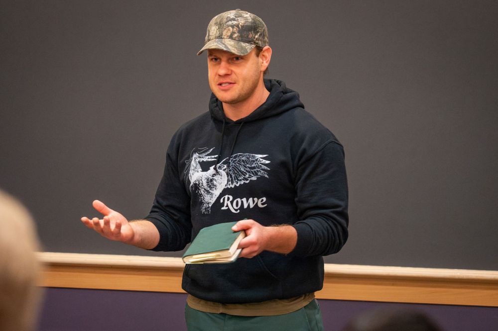 Ethan Tapper, dressed in a black sweatshirt, green pants, and camo baseball cap, holds a book in his hand as he speaks in an auditorium.