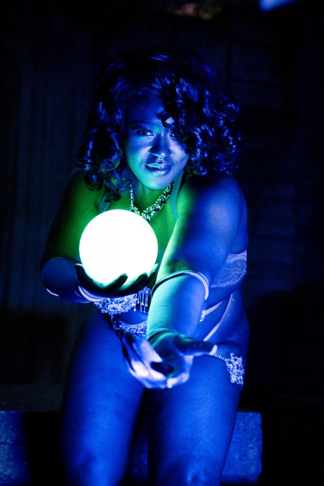 Jue Ve Jones wearing a burlesque costume, holding a glowing orb, sitting in front of an concrete art fountain in the dark, lit by blue light and green from the sphere 
