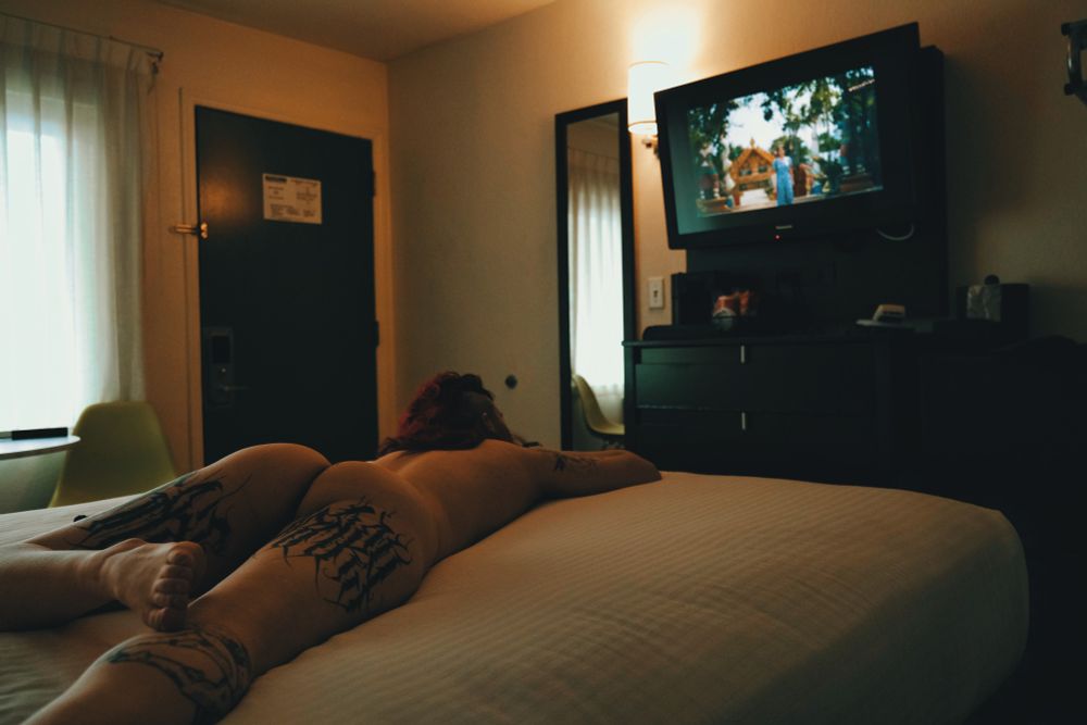 Bex lying naked on the bed watching television in a small hotel room