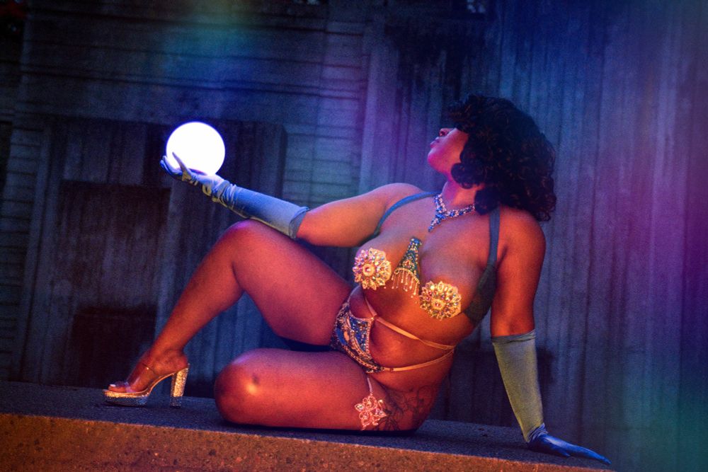 Jue Ve Jones wearing a burlesque costume, holding a glowing orb, sitting in front of an angular concrete art fountain, lit by red right