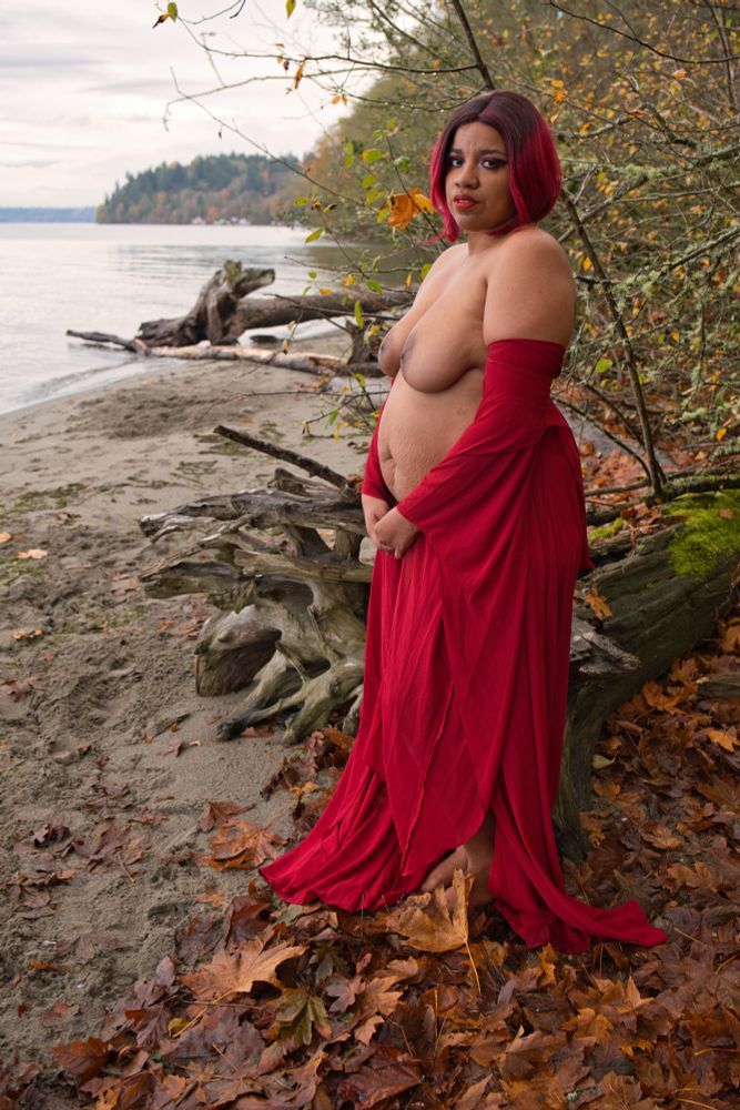 Bella Nazimova stands on a cloudy autumn beach in a red gown