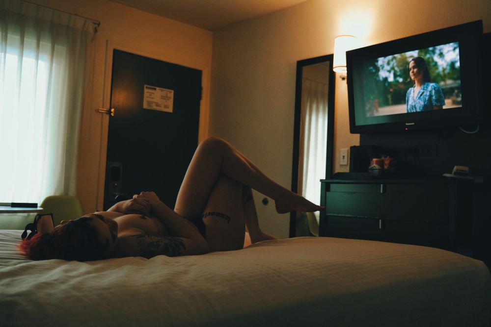 Bex lying naked on the bed watching television in a small hotel room