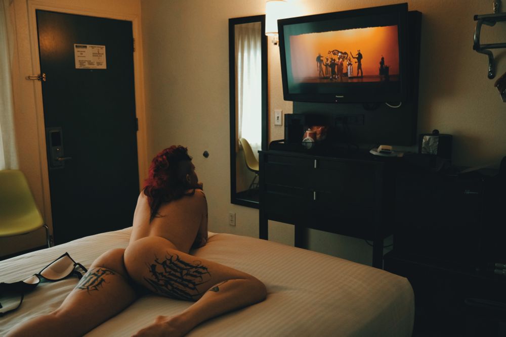 Bex lying naked on the bed watching television in a small hotel room