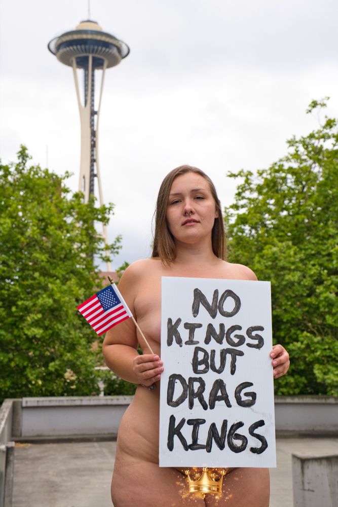 Honey stands nude in front of the Seattle Space Needle holding a protest sign that says "no kings but drag kings" and an American flag (because the flag should still be an anti-fascist symbol)
