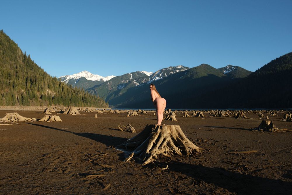 Honey stands nude on a log in a dry lakebed, with mountains in the background, and long shadows stretching behind 