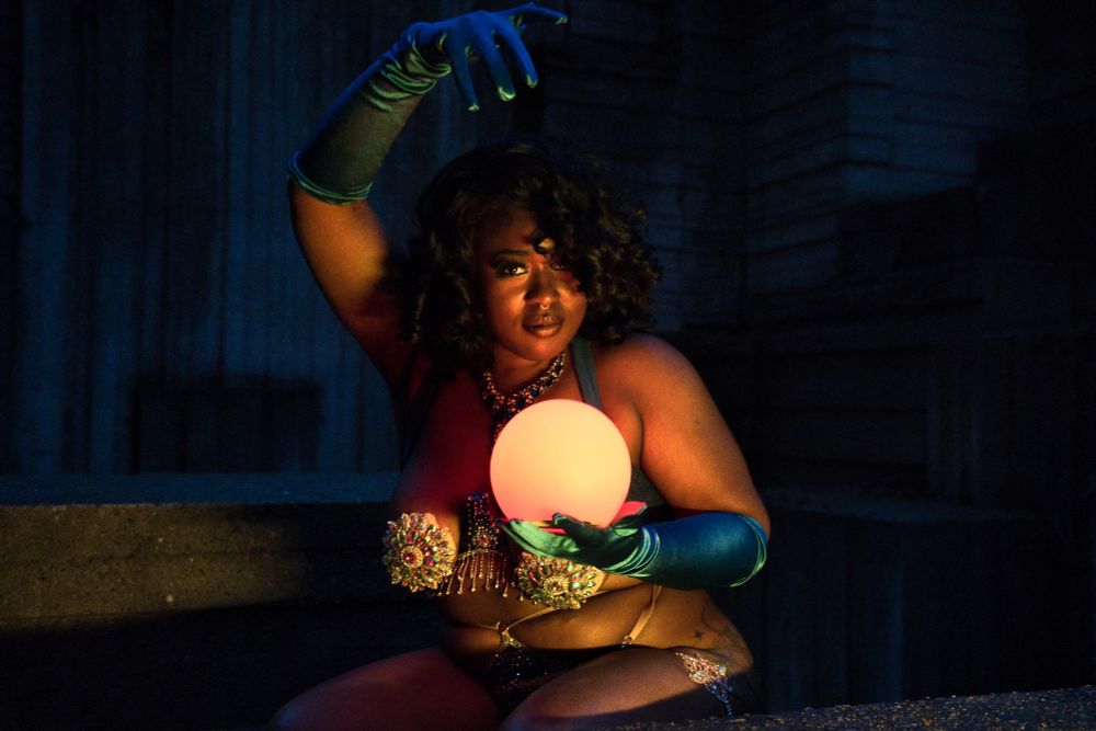 Jue Ve Jones wearing a burlesque costume, holding a glowing orb, sitting in front of an angular concrete art fountain