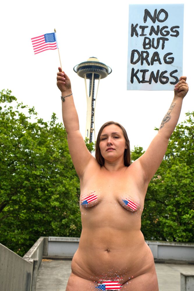 Honey stands naked in front of the Seattle Space Needle holding up a sign that says "No Kings But Drag Kings" and a small American flag