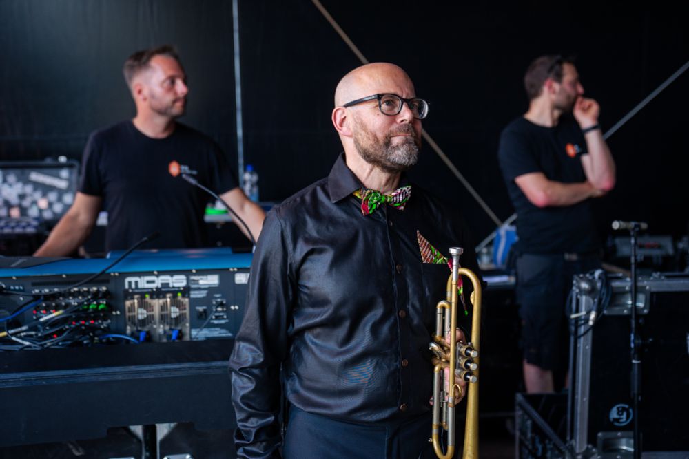 Person holding a trumpet, standing in front of a mixing desk with two sound techs behind it.