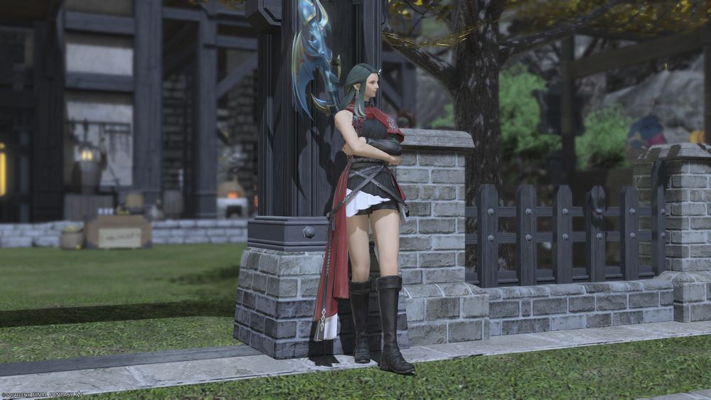 A photo of Xiuying, a light-skinned Elezen with long green hair. She wears a black-and-red tunic with a long tail, black shorts, and knee-high boots. She leans casually against a wall with her arms crossed.
