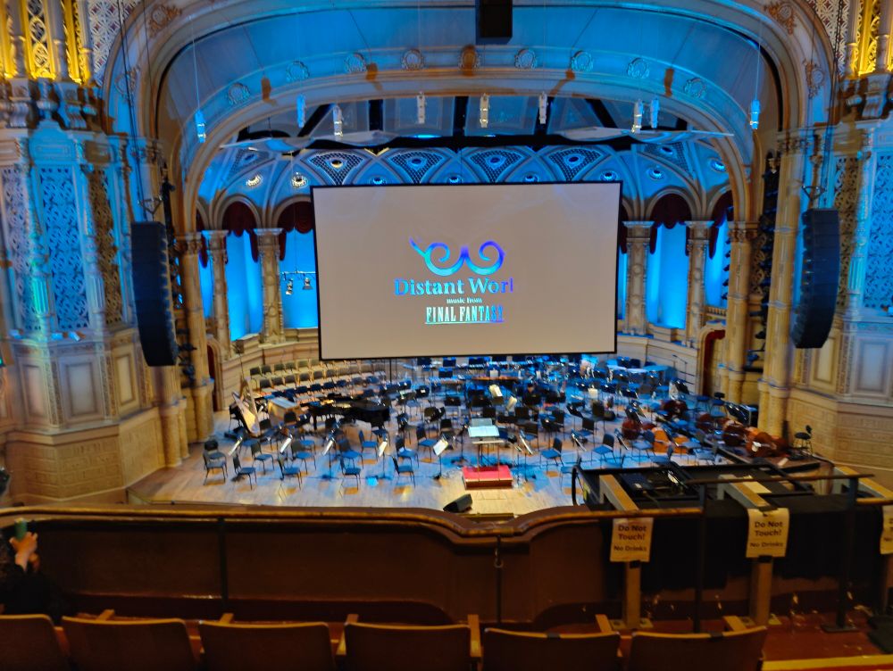 A concert stage for an orchestra, viewed from the upper level. The stage is filled with empty chairs and sheet stands, with a conductor's stand in the front-center. Overhead is an enormous flat screen with the logo "Distant Worlds: Music from Final Fantasy".