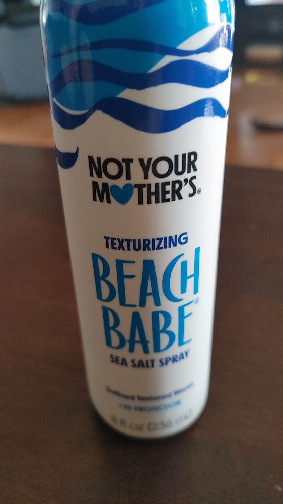 White spray can of Not Your Mother's brand Texturizing Beach Babe Sea Salt Spray. There is a stylizing ocean graphic at the top of the can.